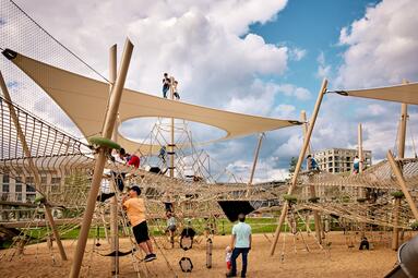 Vergrößerte Ansicht von Ein Spielplatz mit Holzpfählen, dazwischen Netze, darüber ein Sonnensegel. Kinder Spielen in der Spielstation "Vernetzung" in der Parkschale Käfertal, Spinelli-Park, Mannheim