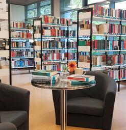 Vergrößerte Ansicht von Ein Foto aus der Stadtteilbibliothek Herzogenried. Auf einem runden, silbernen Beistelltisch liegen Bücher in zwei flachen Stapeln aufeinander. Dazwischen eine Vase mit Blume, dahinter ein Kaffeebecher. Hinter dem Tisch stehen zwei dunkelgraue Stoffsessel.