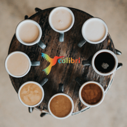 Vergrößerte Ansicht von Kaffeetassen stehen im Kreis auf einem runden Tisch. In der Mitte ein bunter Colibri. Es ist das Logo des "Café Colibri".