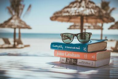 Vergrößerte Ansicht von Eine Sonnenbrille auf einem Stapel Bücher. Dahinter Strand, Sonnenschirme, Liegen und das Meer.