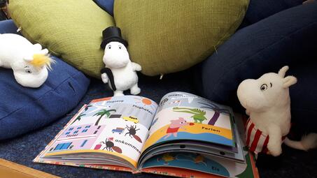 Vergrößerte Ansicht von Ein aufgeschlagenes Bilderbuch und drum herum mehrere Kissen und Mumin-Stofftiere