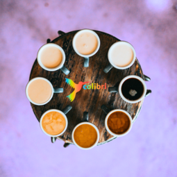 Vergrößerte Ansicht von Ein Blick von oben auf einen runden Tisch. Darauf stehen Tassen im Kreis, gefüllt mit unterschiedlich hellem und dunklem Kaffee. Der Hintergrund ist lila. In der Mitte ein bunter Kolibri-Vogel neben dem Wort colibri.