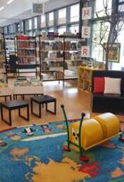 Vergrößerte Ansicht von ein Foto vom Bilderbuchbereich der Bibliothek. Man sieht Regale, Hocker, einen bunten Teppich und Dekoration