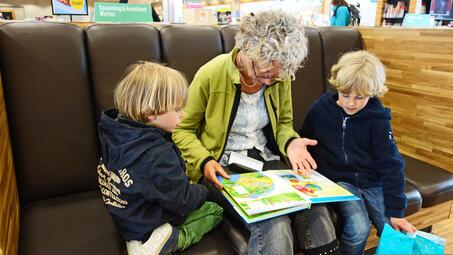 Vergrößerte Ansicht von Eine Frau und zwei Kinder sitzen auf einem Sofa. Die Frau liest den Kindern ein Buch vor.