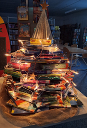 Vergrößerte Ansicht von eine auf einem Tisch stehende Nachbildung eines Tannenbaums, die aus übereinander geschichteten Büchern und einer Lichterkette besteht.