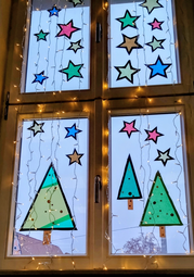 Vergrößerte Ansicht von Ein Fenster, an dem gebastelte Tannenbäume und Sterne kleben und an dem eine Lichterkette hängt.
