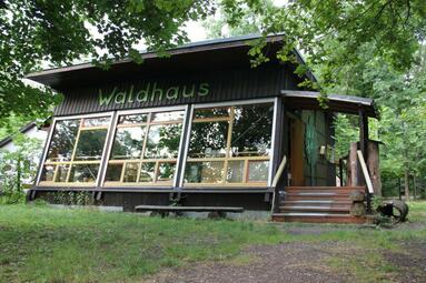 Vergrößerte Ansicht von Einstöckige Hütte mit großer Fensterfront, darüber der Schriftzug "Waldhaus"